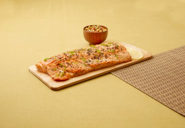 Baked Salmon on a Cedar Plank with a Bowl of Corn Salsa on a Yellow Surface and Brown Placemat
