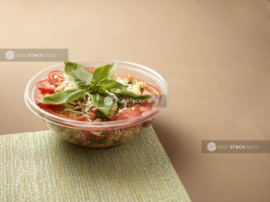 Take-out Container of Pasta Salad with Sliced Tomatoes and Fresh Basil Leaves on a Placemat - Variation