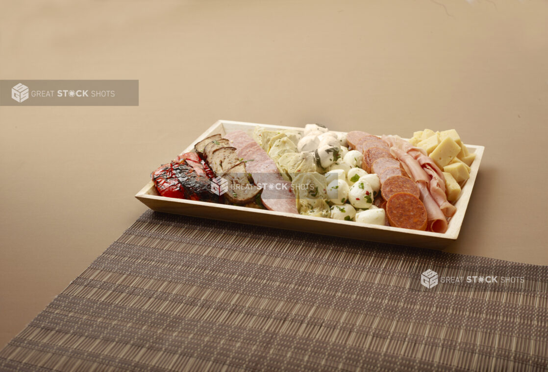 A Rectangular Wood Tray of Antipasto with Assorted Cheese, Deli Meats and Vegetables on a Brown Surface and Placemat