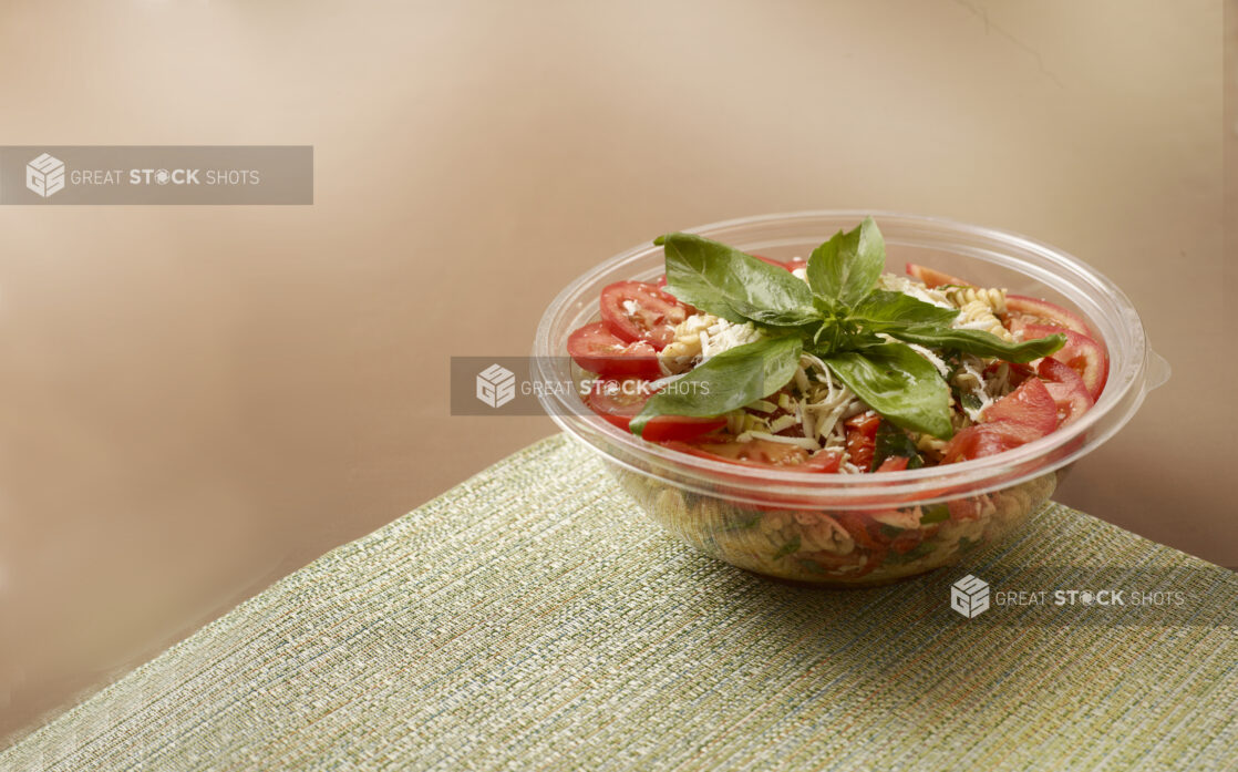 Take-out Container of Pasta Salad with Sliced Tomatoes and Fresh Basil Leaves on a Placemat