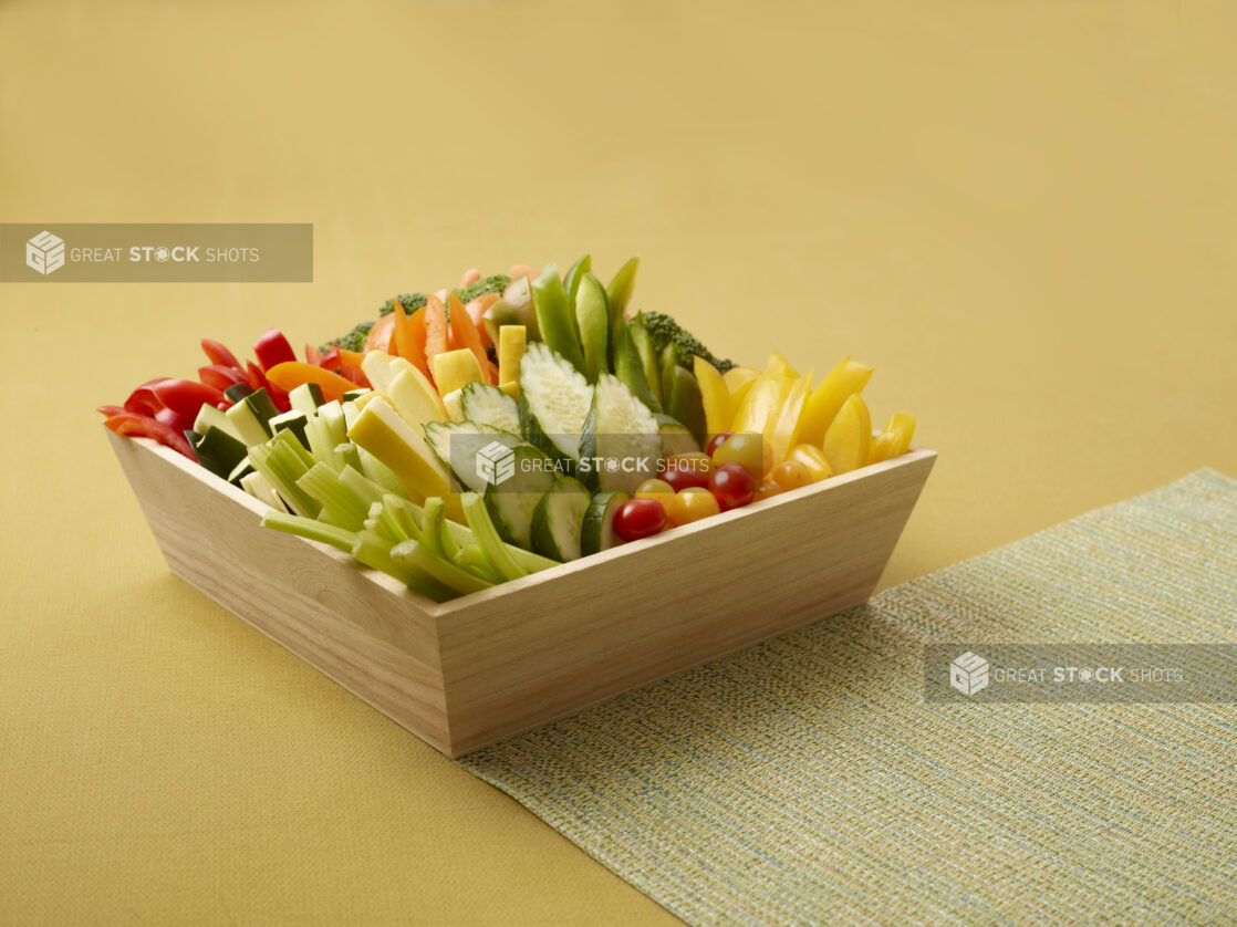 Assorted Fresh Vegetable Sticks in a Deep Wooden Tray on a Yellow Background