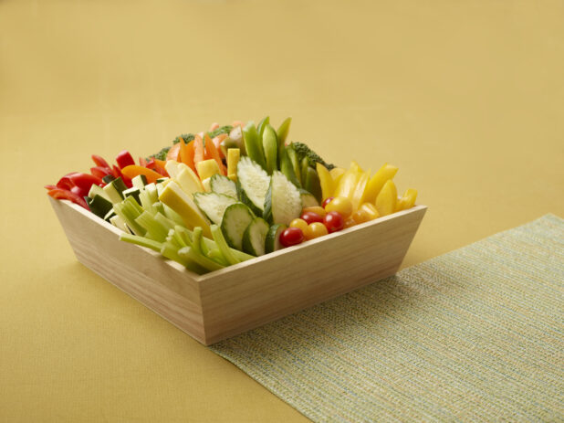 Assorted Fresh Vegetable Sticks in a Deep Wooden Tray on a Yellow Background