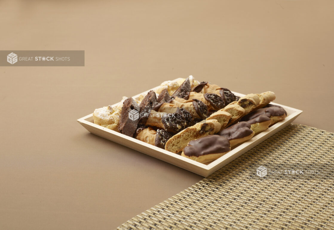 Square Wood Tray with Assorted Italian Desserts - Biscottis, Cannoli and Mini Eclairs on a Brown Surface and Placemat - for Catering