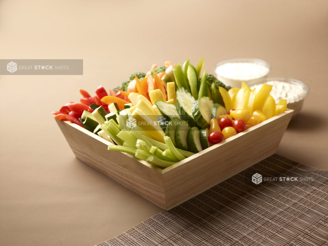 Assorted Fresh Vegetable Sticks in a Deep Wooden Tray on a Brown Background with Placemat and Creamy Dipping Sauces