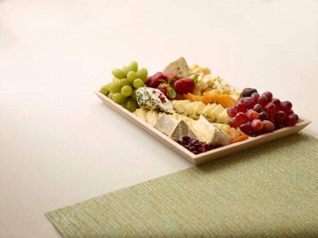 Hors D'Oeuvre Cheese Platter with Assorted Sliced Cheese, Dried Fruits and Fresh Grapes and Strawberries on a Beige Surface and Green Placemat - for Catering