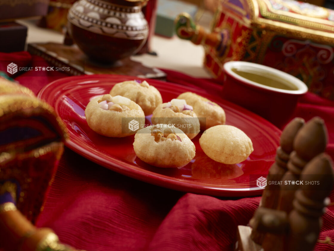 Panipuri - Indian Wheat Shells - Filled with Chickpeas and Red Onions on a Red Ceramic Dish with Green Chilli Water on a Red Tablecloth