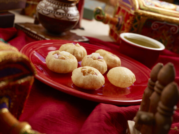 Panipuri - Indian Wheat Shells - Filled with Chickpeas and Red Onions on a Red Ceramic Dish with Green Chilli Water on a Red Tablecloth