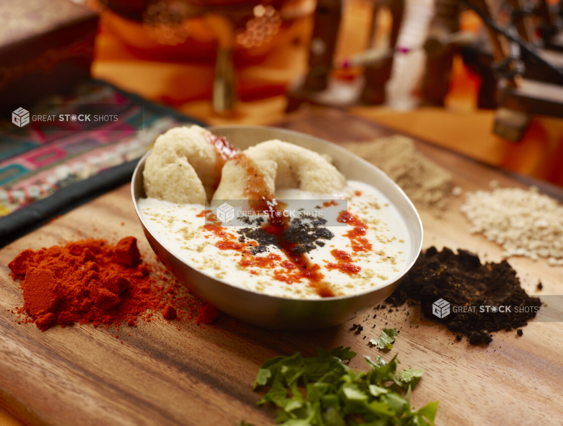 Raita Indian Yogurt Dish with Medu Vada Lentil Doughnuts in a Ceramic Bowl Surrounded by Spices and Ingredients on a Wooden Cutting Board