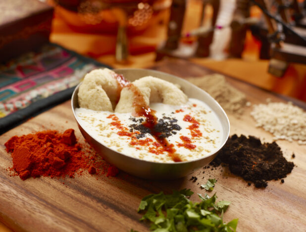 Raita Indian Yogurt Dish with Medu Vada Lentil Doughnuts in a Ceramic Bowl Surrounded by Spices and Ingredients on a Wooden Cutting Board