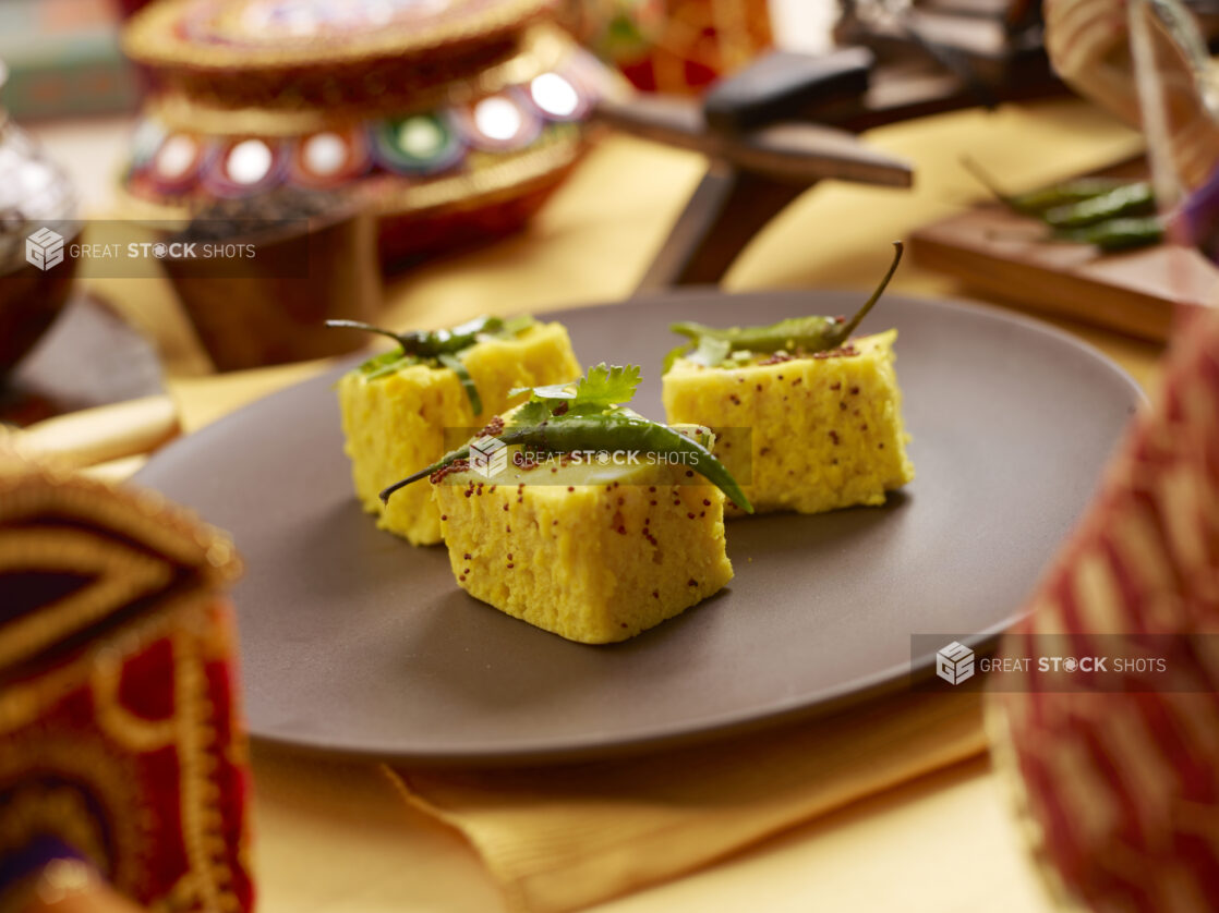 Khaman Dhokla - Indian Steamed Chickpea Flour Cakes - on a Brown Ceramic Dish with Yellow Placemats and Tablecloth