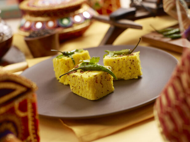Khaman Dhokla - Indian Steamed Chickpea Flour Cakes - on a Brown Ceramic Dish with Yellow Placemats and Tablecloth