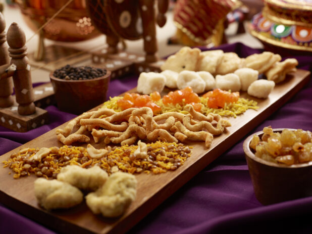 An Assortment of Indian Sweets - Mithai, Boondi, Bombay Mix, on a Wooden Platter with Indian Decorative Pieces on a Table in an Indoor Setting