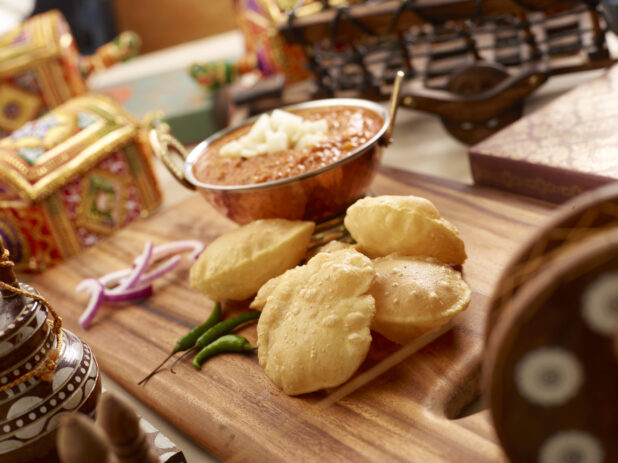 Copper Bowl of Puri Bhaji – Deep Fried Puff Bread and Thick Vegetable Curry – with Fresh Ingredients on a Wooden Platter in an Indoor Setting