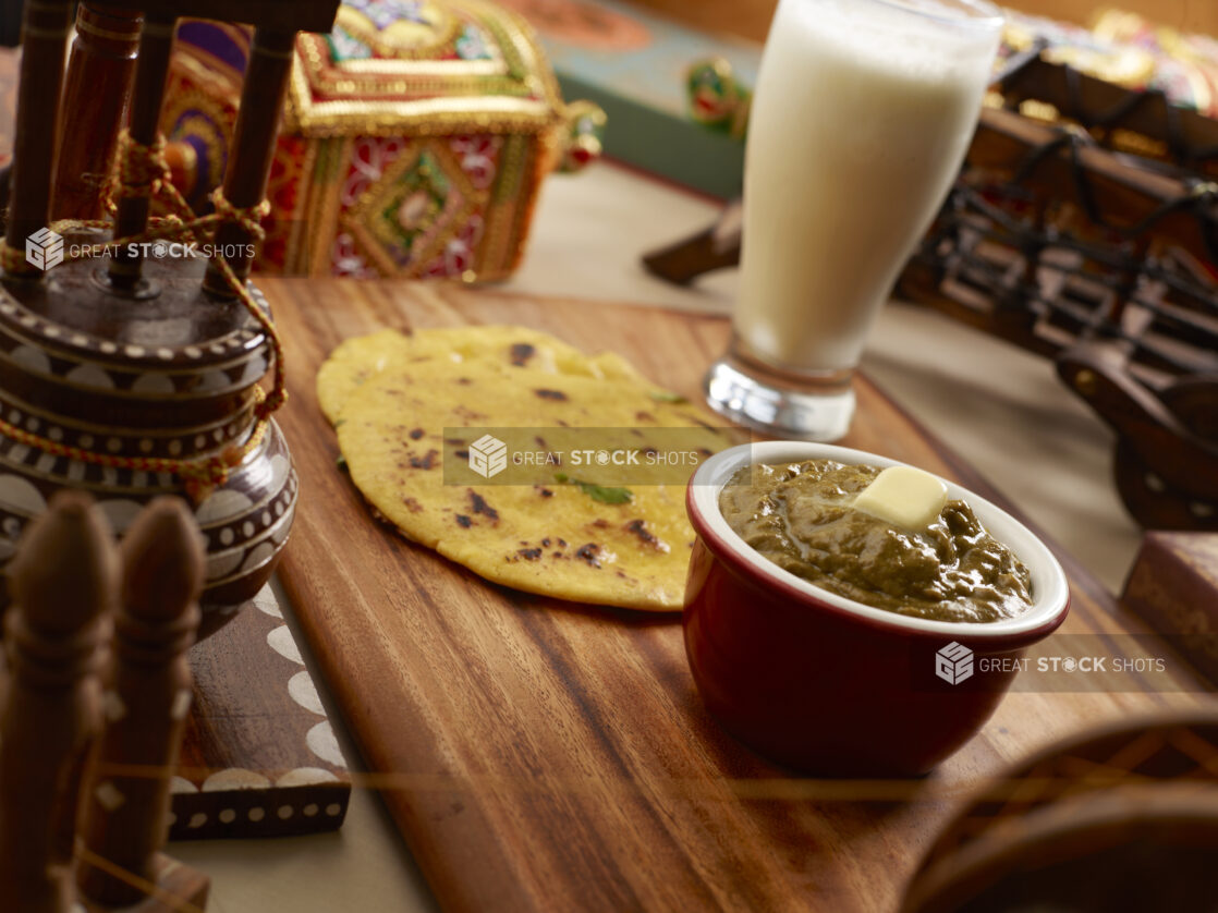 An Indian Combo Meal of Aloo Kulcha Bread, Saag Curry with Butter and a Glass of Lassi on a Wooden Platter in an Indoor Setting