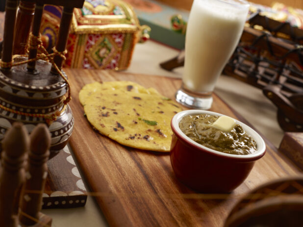 An Indian Combo Meal of Aloo Kulcha Bread, Saag Curry with Butter and a Glass of Lassi on a Wooden Platter in an Indoor Setting