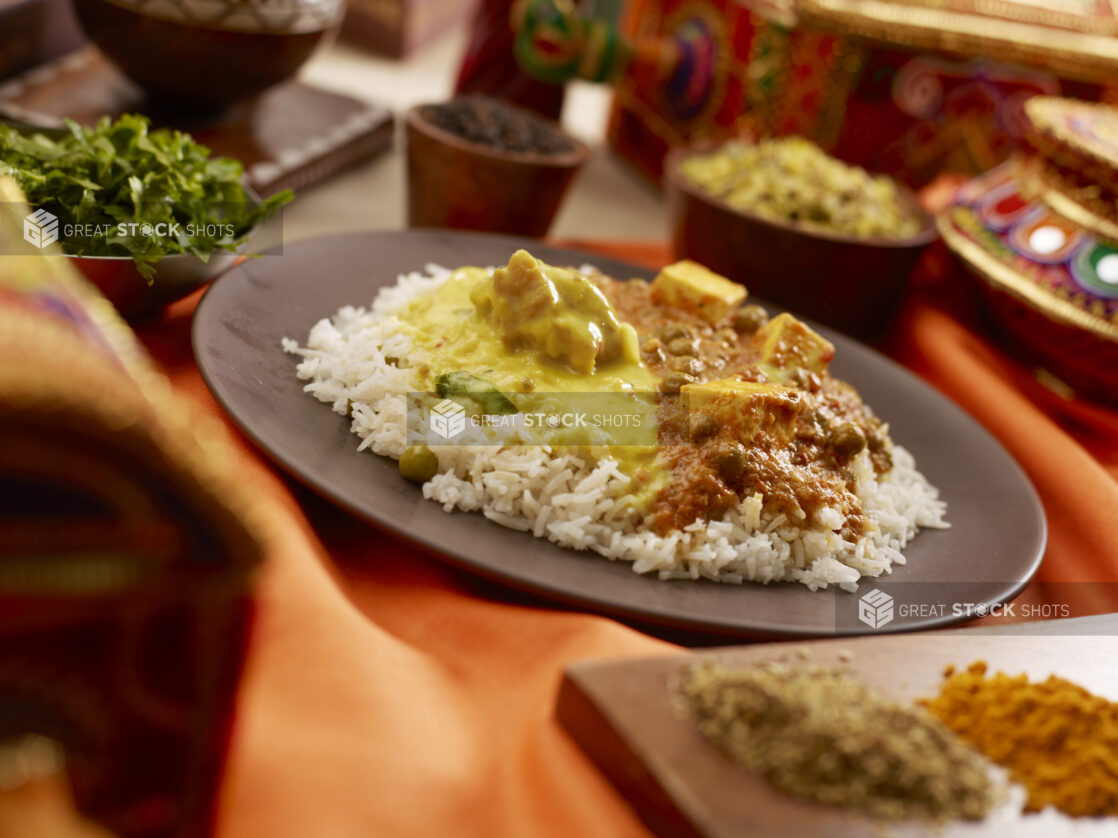 Two Types of Curry - Vegetable Korma and Paneer Daal Makhani - Over Basmati Rice Surrounded by Spices and Toppings on an Orange Tablecloth