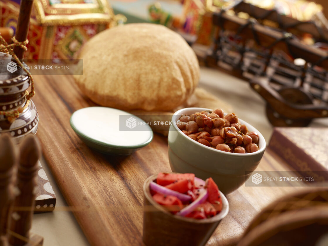 Indian Breakfast of Channa Bhatura – Spiced Chickpeas and Indian Puff Bread with Side Dishes of Raita and Pickled Vegetables on a Wooden Tray