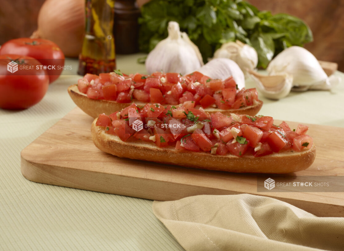 Two Toasted Italian Bread Slices Topped with Fresh Tomato Bruschetta on a Wooden Cutting Board with Fresh Vegetable Ingredients in Background in an Indoor Setting