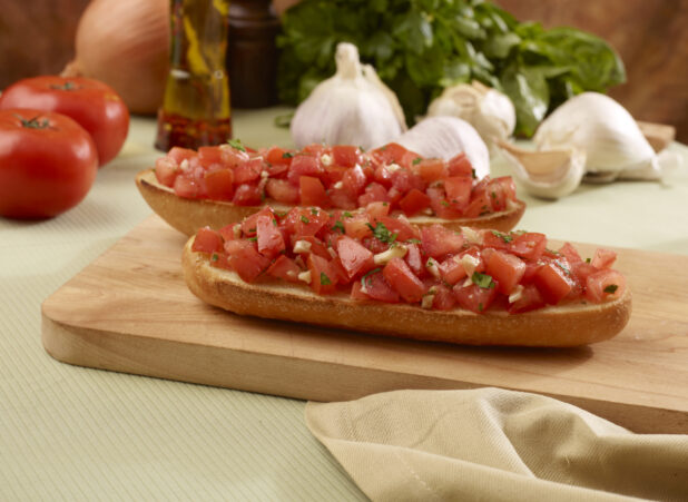 Two Toasted Italian Bread Slices Topped with Fresh Tomato Bruschetta on a Wooden Cutting Board with Fresh Vegetable Ingredients in Background in an Indoor Setting