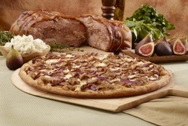 Specialty Pizza with Porchetta, Red Onions, Feta Cheese and Fig Sauce on a Wooden Pizza Paddle with Whole Porchetta, Figs, Fresh Herbs and Feta Cheese in the Background