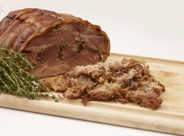 Close Up of Italian Porchetta Roast and Shaved Pork Pieces on a Wooden Cutting Board with Fresh Herbs Isolated on a White Background