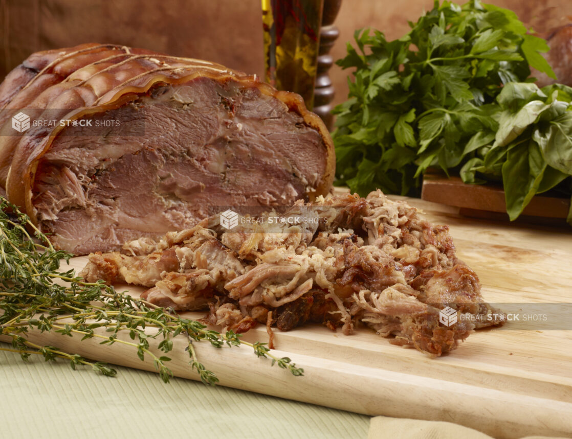 Close Up of Italian Porchetta Roast and Shaved Pork Pieces on a Wooden Cutting Board with Fresh Herbs on a Green Table Cloth Surface in an Indoor Setting