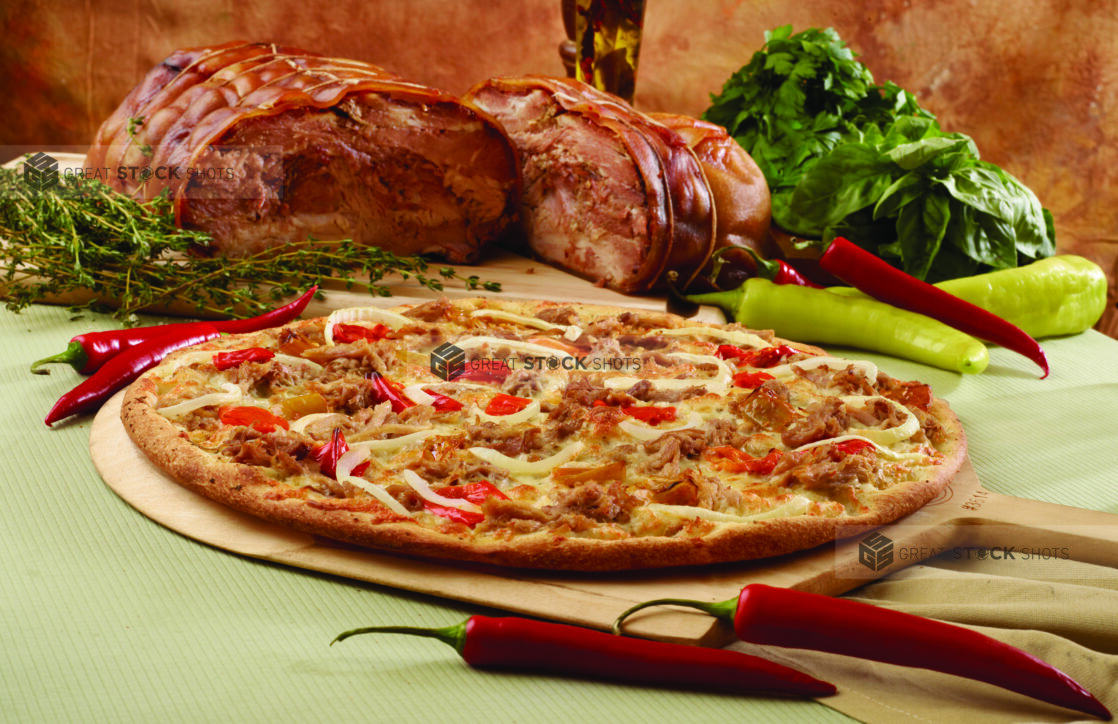 Specialty Pizza with Porchetta, Sliced White Onions and Hot Banana Peppers on a Wooden Pizza Paddle with Whole Porchetta, Green and Red Banana Peppers and Fresh Herbs in the Background