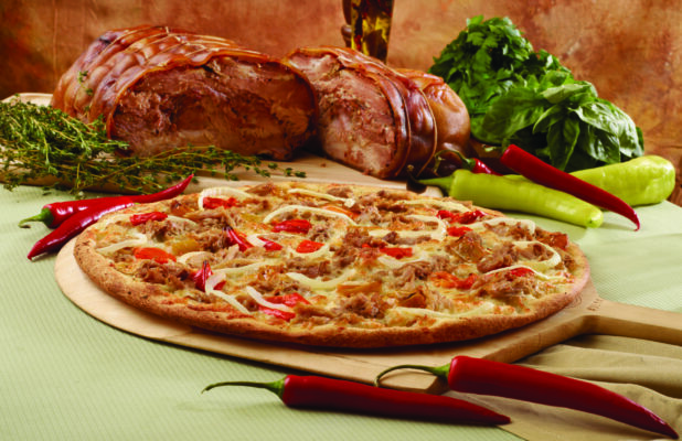 Specialty Pizza with Porchetta, Sliced White Onions and Hot Banana Peppers on a Wooden Pizza Paddle with Whole Porchetta, Green and Red Banana Peppers and Fresh Herbs in the Background