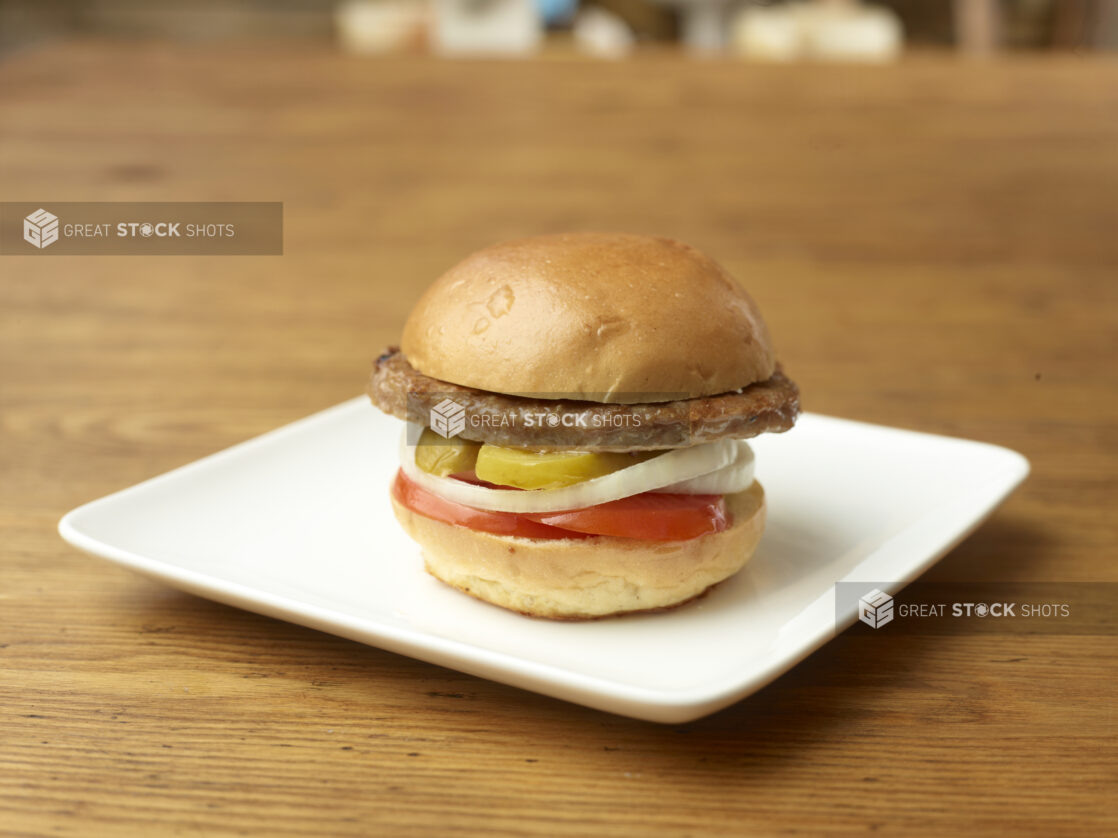 Single Patty Flame-Broiled Hamburger with Sliced Onions, Tomatoes and Pickles on a Square White Ceramic Dish on a Rustic Wood Surface