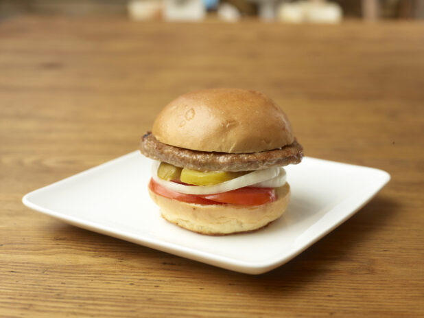 Single Patty Flame-Broiled Hamburger with Sliced Onions, Tomatoes and Pickles on a Square White Ceramic Dish on a Rustic Wood Surface