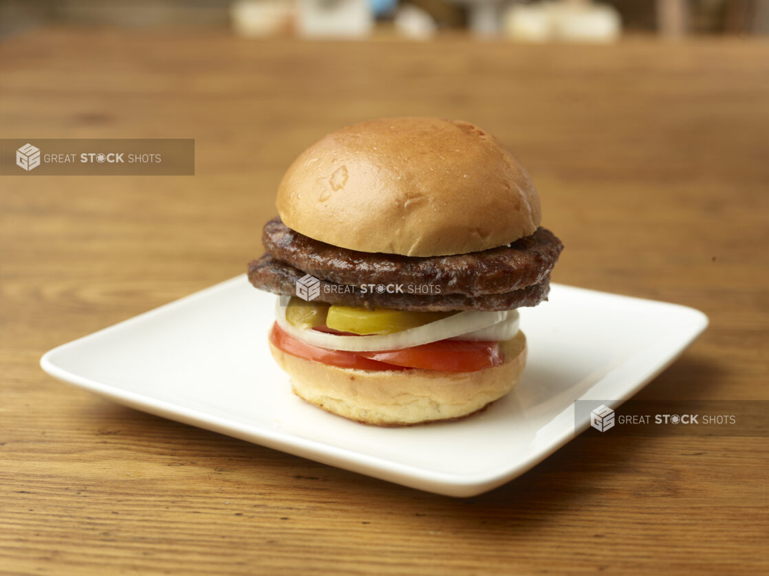 Double All Beef Patty Flame-Broiled Hamburger with Sliced Onions, Tomatoes and Pickles on a Square White Ceramic Dish on a Rustic Wood Surface