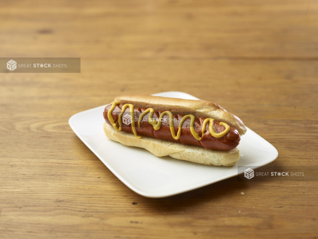 Plain Hot Dog with Yellow Mustard on a Rectangular White Ceramic Dish on a Rustic Wooden Table