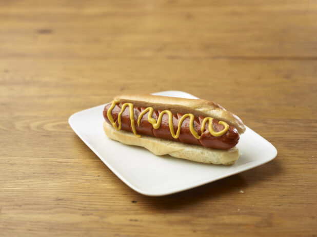 Plain Hot Dog with Yellow Mustard on a Rectangular White Ceramic Dish on a Rustic Wooden Table