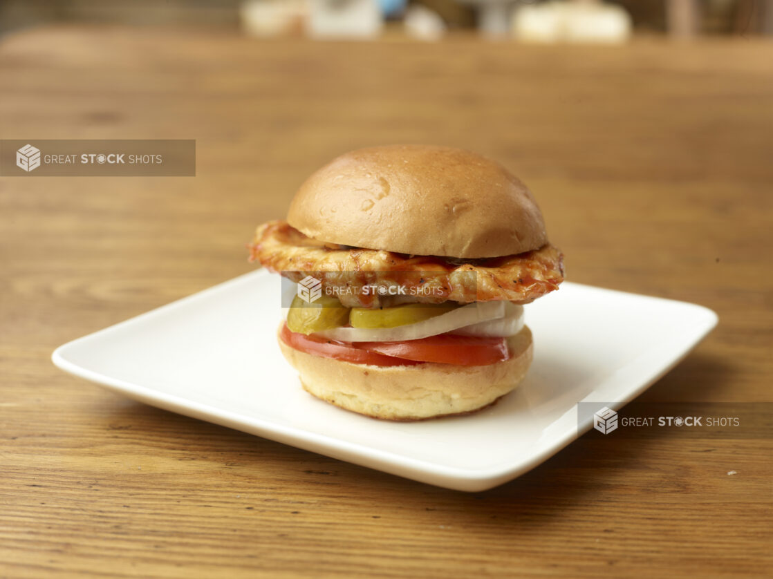 Grilled Barbecue Chicken Burger with Sliced Pickles, White Onions and Tomatoes on a Square White Ceramic Plate on a Rustic Wood Surface