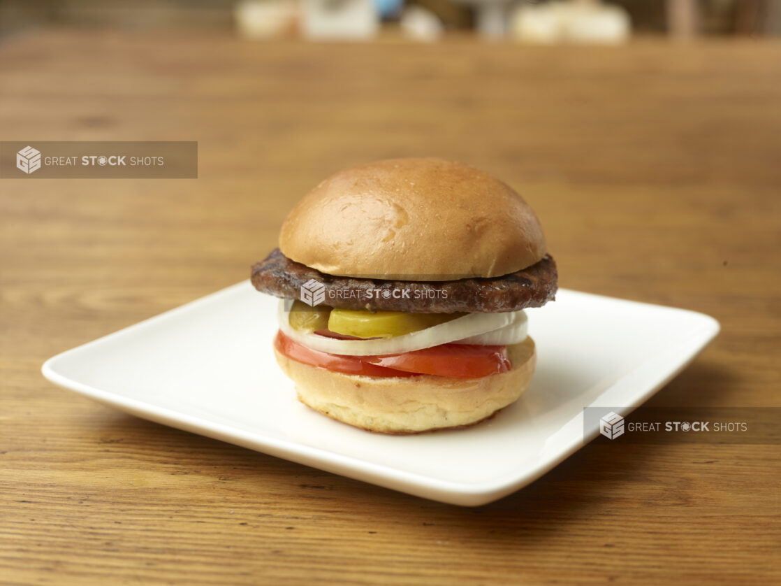 Single All Beef Patty Flame-Broiled Hamburger with Sliced Onions, Tomatoes and Pickles on a Square White Ceramic Dish on a Rustic Wood Surface