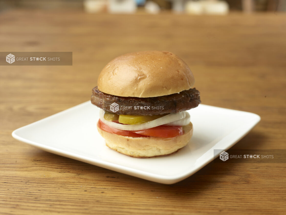 Single Plant-Based Patty Flame-Broiled Hamburger with Sliced Onions, Tomatoes and Pickles on a Square White Ceramic Dish on a Rustic Wood Surface