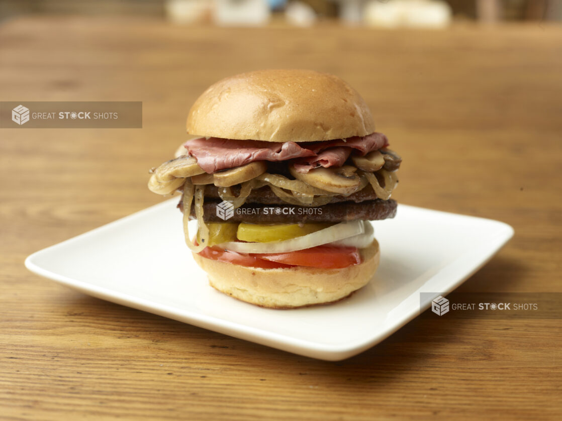 Gourmet Double Burger with Pastrami, Sautéed Mushrooms and Onions, Sliced Pickles, Onions and Tomatoes on a Square White Dish on a Rustic Wooden Surface