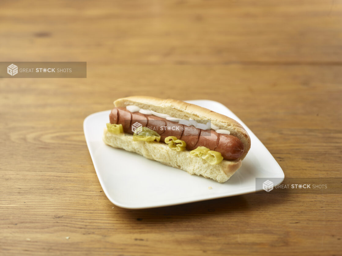 Hot Dog with Hot Banana Peppers and White Onion Slices on a Rectangular White Ceramic Dish on a Rustic Wooden Table - Variation