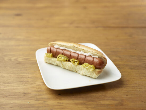 Hot Dog with Hot Banana Peppers and White Onion Slices on a Rectangular White Ceramic Dish on a Rustic Wooden Table - Variation