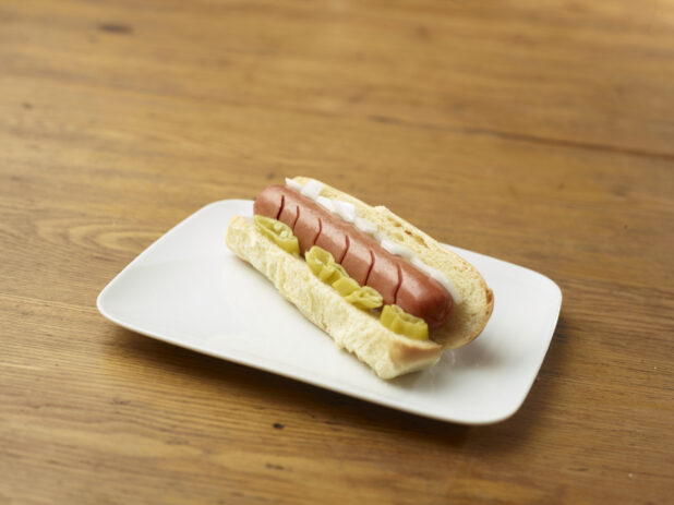 Hot Dog with Hot Banana Peppers and White Onion Slices on a Rectangular White Ceramic Dish on a Rustic Wooden Table