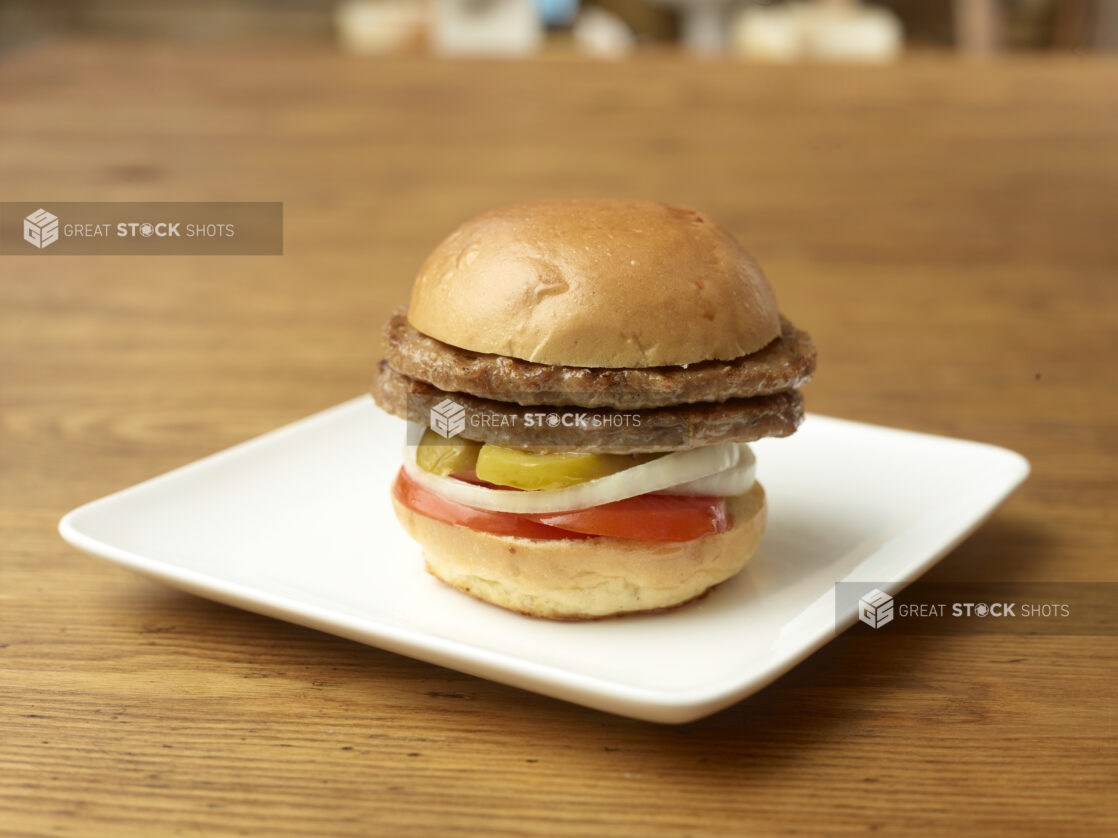 Double Patty Flame-Broiled Hamburger with Sliced Onions, Tomatoes and Pickles on a Square White Ceramic Dish on a Rustic Wood Surface