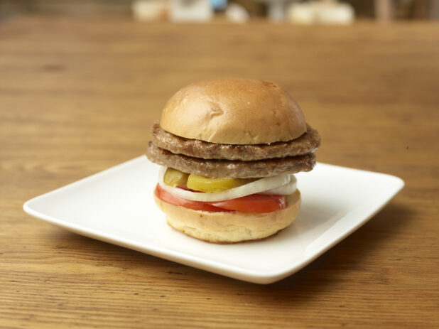 Double Patty Flame-Broiled Hamburger with Sliced Onions, Tomatoes and Pickles on a Square White Ceramic Dish on a Rustic Wood Surface