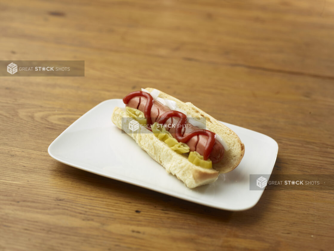 Hot Dog with Ketchup, Hot Banana Peppers and White Onion Slices on a Rectangular White Ceramic Dish on a Rustic Wooden Table