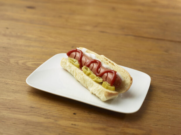 Hot Dog with Ketchup, Hot Banana Peppers and White Onion Slices on a Rectangular White Ceramic Dish on a Rustic Wooden Table