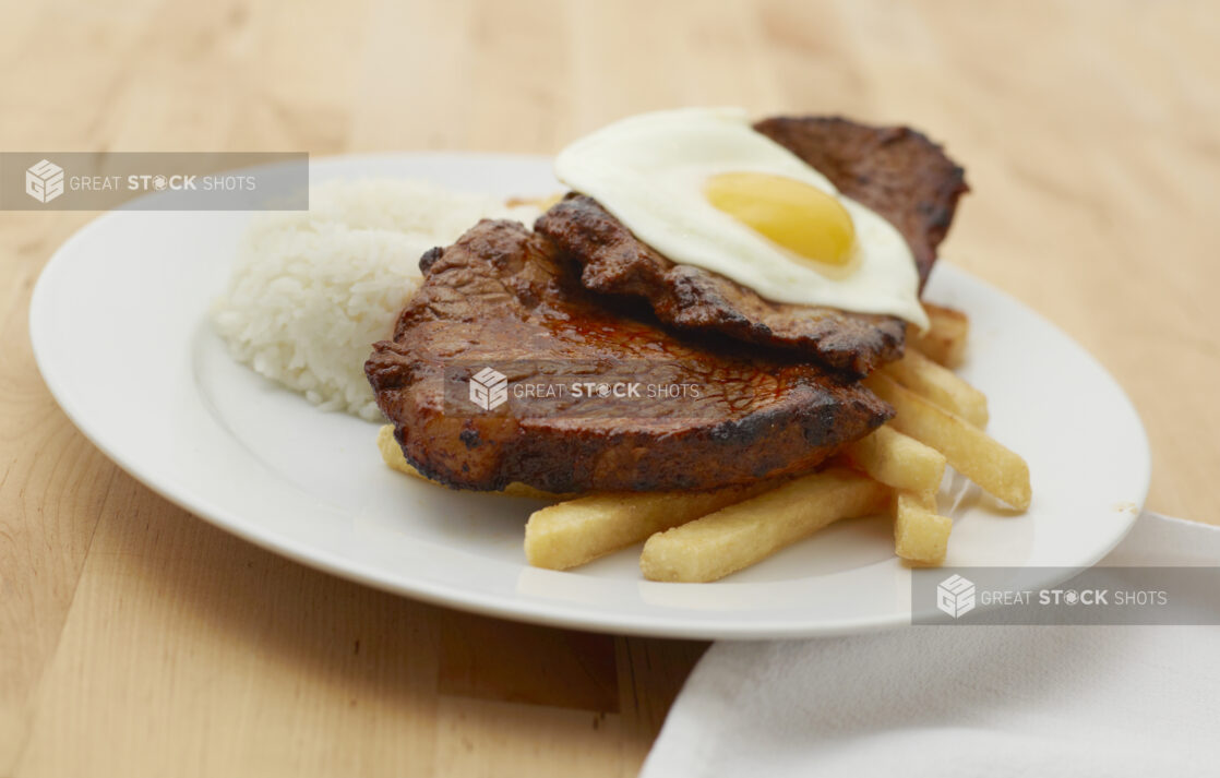 Steak, Sunny Side Up Egg, French Fries and White Rice on a Round White Ceramic Dish on a Light Wooden Table