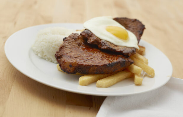Steak, Sunny Side Up Egg, French Fries and White Rice on a Round White Ceramic Dish on a Light Wooden Table