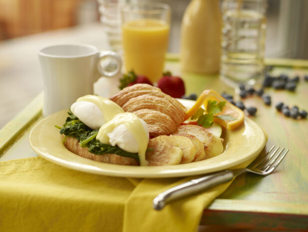 Breakfast Platter with a Spinach Eggs Benedict Croissant Sandwich, Home Fries and Fresh Fruit with other Breakfast Items on a Painted Wooden Tray in an Indoor Setting