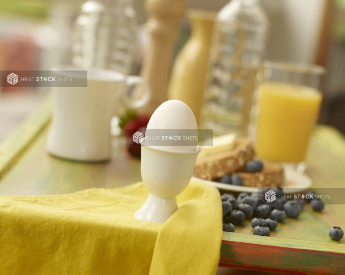 Boiled Egg in a White Ceramic Egg Cup with Breakfast Items and Ingredients in the Background on a Painted Wooden Tray