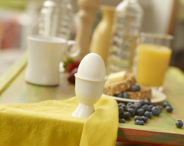 Boiled Egg in a White Ceramic Egg Cup with Breakfast Items and Ingredients in the Background on a Painted Wooden Tray