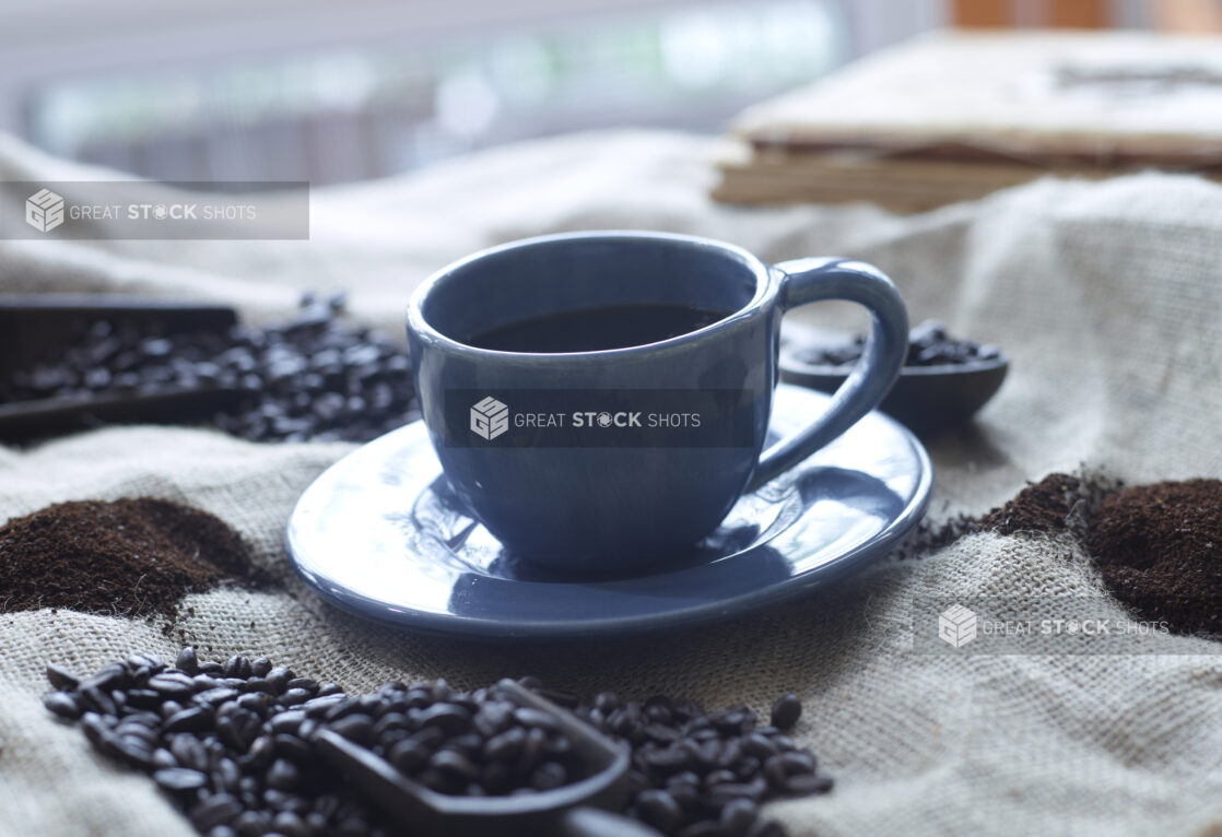 Black Coffee in a Blue Cup with Saucer on a Burlap Material Surface with Coffee Beans and Ground Coffee in an Indoor Setting